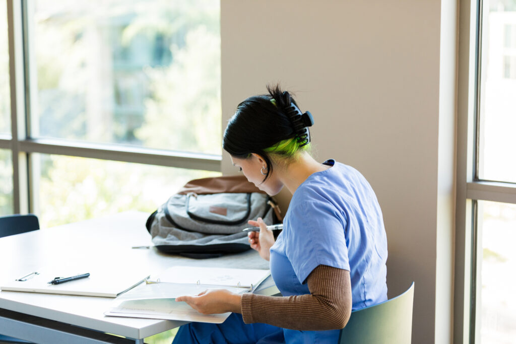 A young woman sits at a desk reviewing paperwork. Natural light streams from a large window.